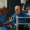 A Black female nurse in blue scrubs with dreadlocks and glasses writes on a clipboard while talking to an older white male patient with glasses who is sitting and holding onto a walker. The discussion could involve medical instructions, such as where to buy Suprep Bowel Prep Kit for a procedure.