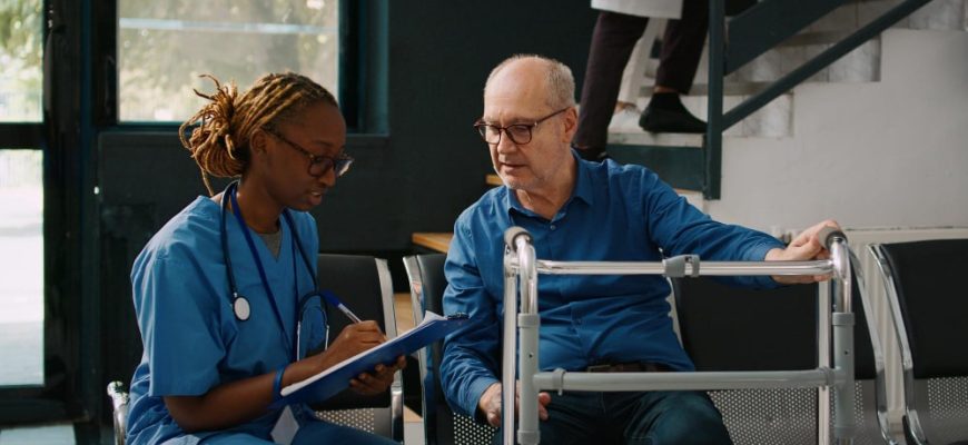 A Black female nurse in blue scrubs with dreadlocks and glasses writes on a clipboard while talking to an older white male patient with glasses who is sitting and holding onto a walker. The discussion could involve medical instructions, such as where to buy Suprep Bowel Prep Kit for a procedure.
